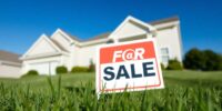 House with for sale sign amidst green grass and sky.